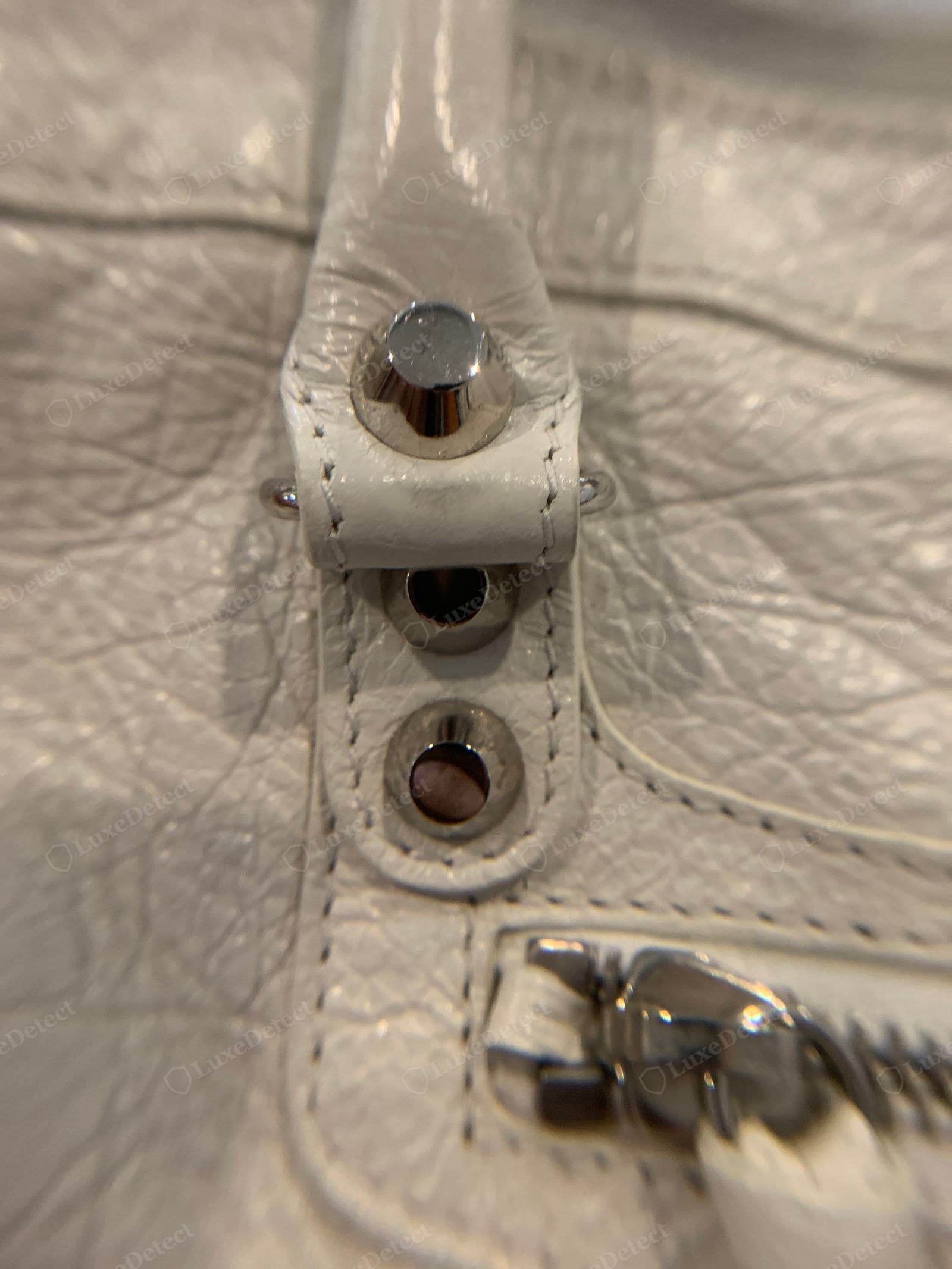 Genuine Balenciaga City hardware — a macro of white distressed leather with smooth domed silver studs, the buckle post, and a ridged Lampo-style zip pull, all one consistent finish