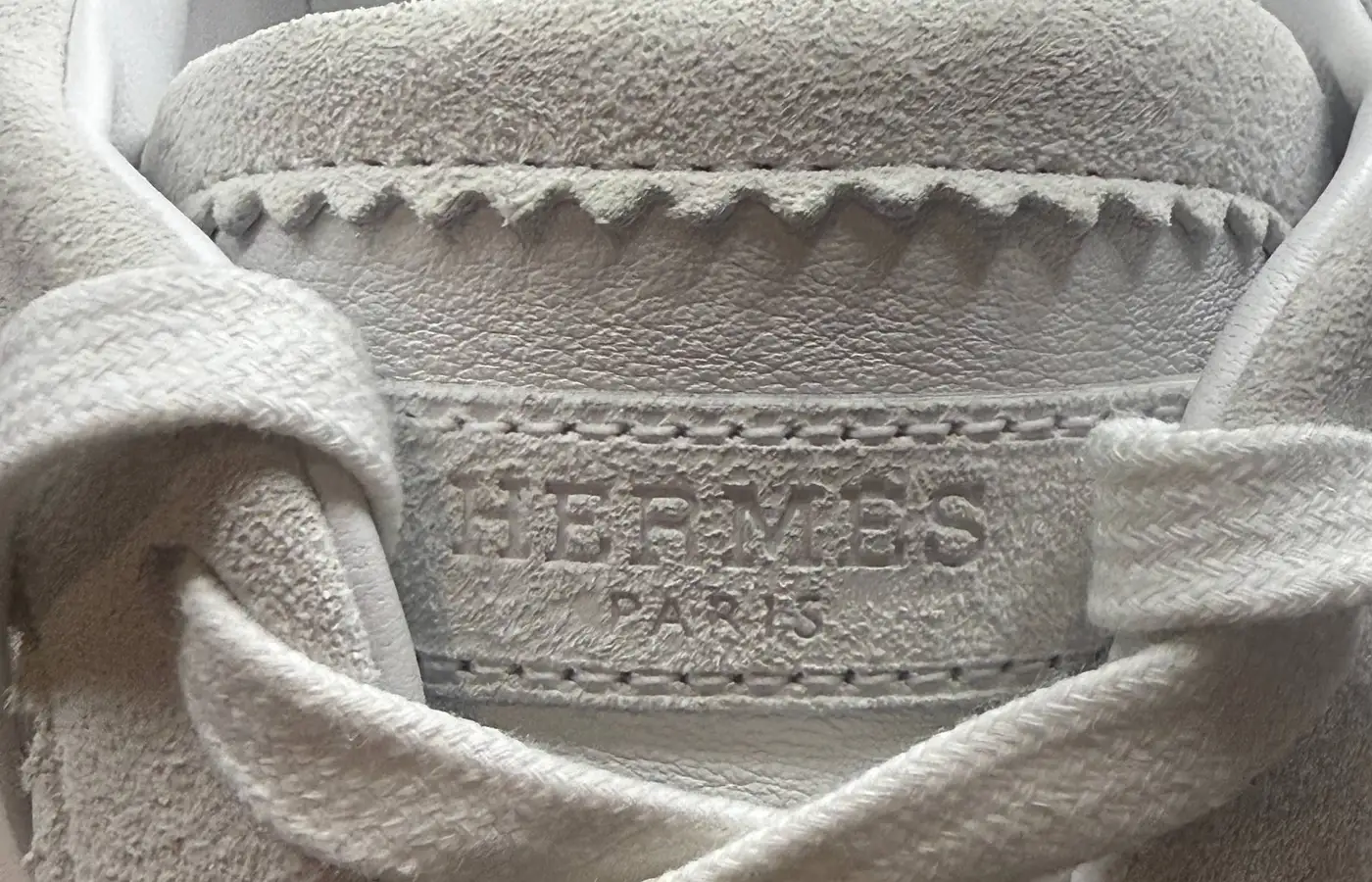 Tongue detail on a white pair — "HERMÈS PARIS" debossed into suede, the tab edges cut in a fine pinked zigzag, and the collar finished with hand whipstitching