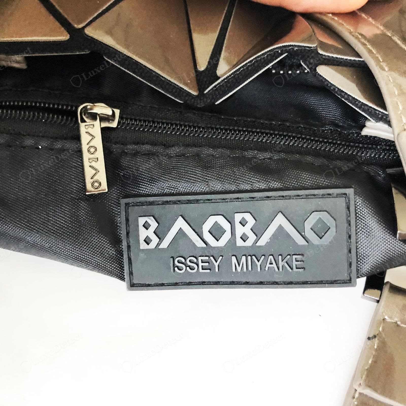 Counterfeit BAOBAO interior — a thin plain black lining, a stuck-on rubber "BAOBAO ISSEY MIYAKE" patch, and a flimsy zip pull with the name stamped vertically down a thin tab