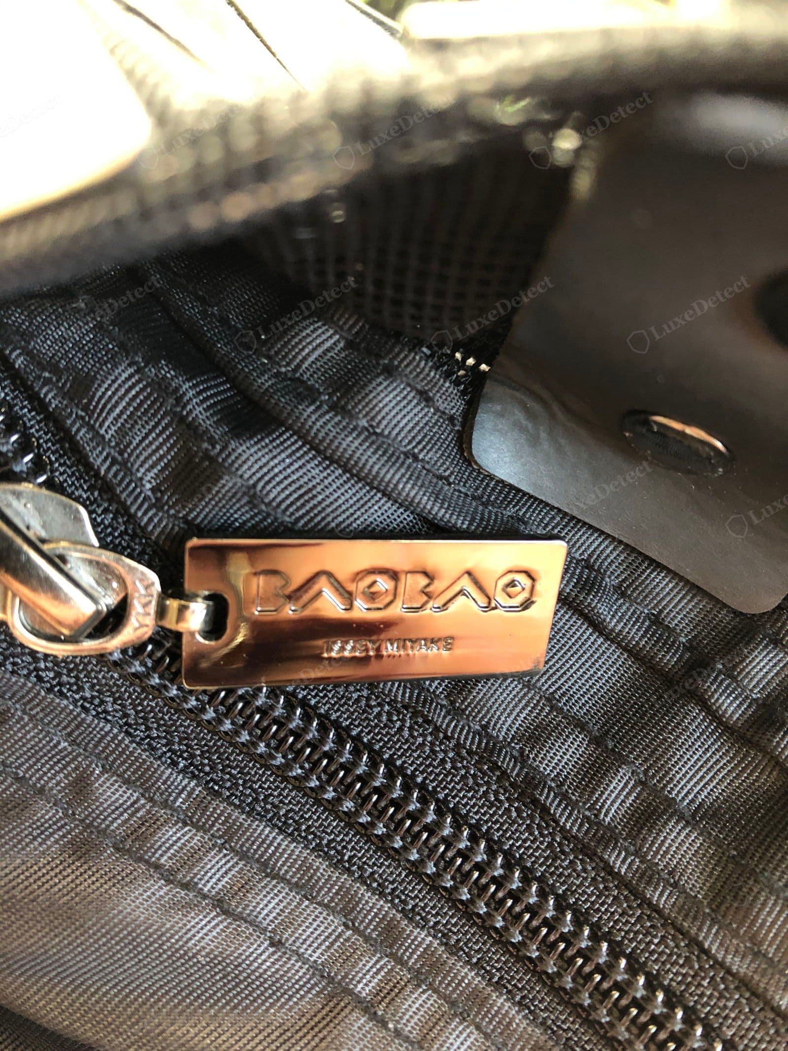 Genuine BAOBAO zip pull — a polished chrome plate with a deep, crisp "BAOBAO / ISSEY MIYAKE" emboss in the geometric typeface, on a YKK-stamped slider, against the branded logo-jacquard lining