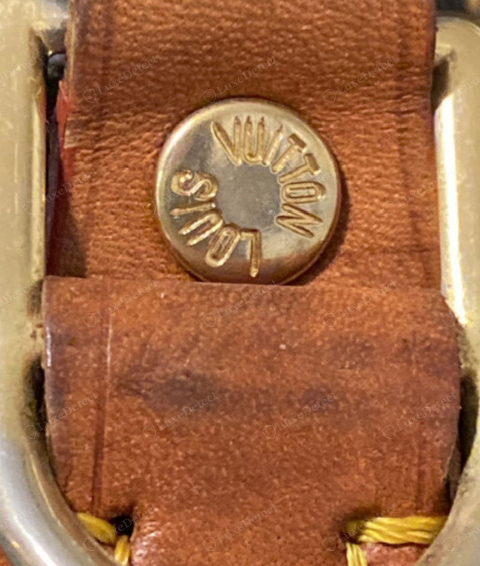 Counterfeit LV Alma trim leather — a strap tab with a "LOUIS VUITTON" engraved rivet, the vachetta blotchy and over-dark orange-brown from a fake aged finish, the grain coarse and the edge rough