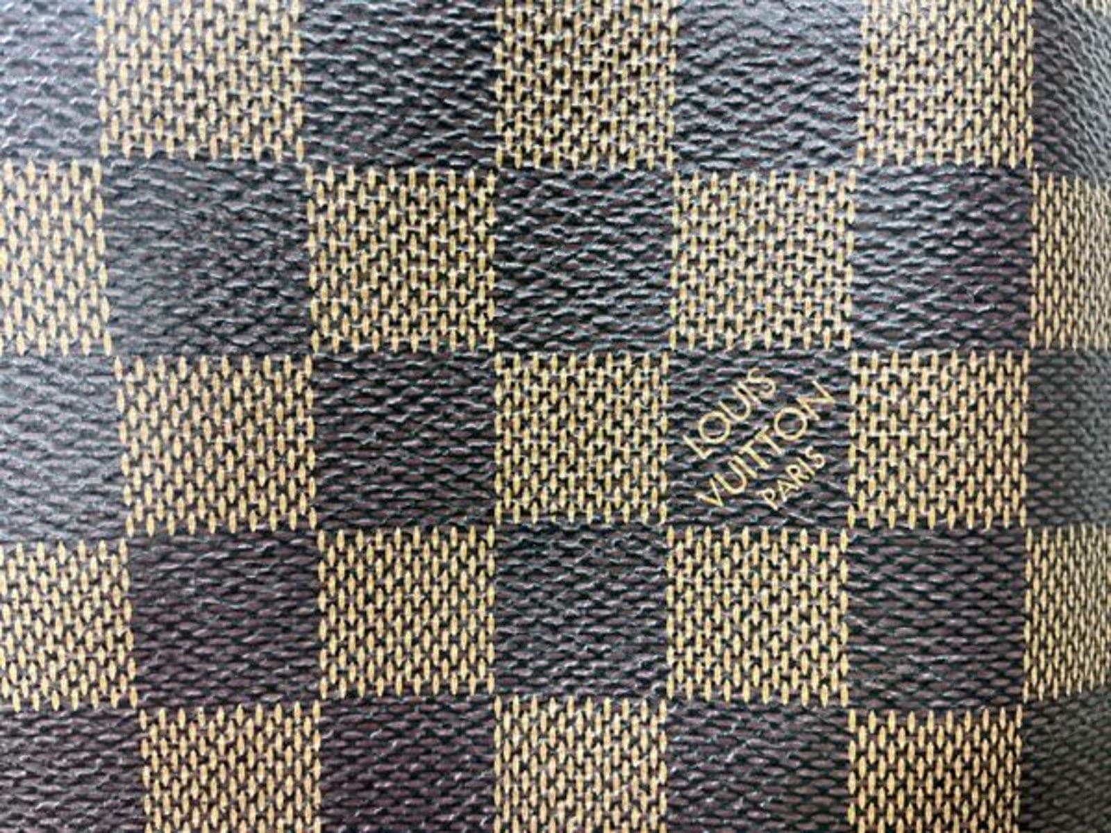 Counterfeit LV canvas (Damier Ebene shown) — the "Louis Vuitton Paris" stamp in its pale square is thin and slightly ragged, and the checkerboard has an over-pronounced woven basketweave texture rather than the smooth coated finish of the genuine canvas