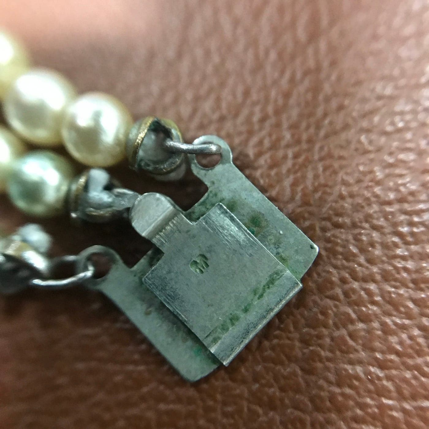 Counterfeit Mikimoto three-strand necklace clasp, reverse. The green verdigris crust across the base-metal clasp is copper-alloy corrosion — sterling silver and gold cannot do this. The maker's stamp at right is a shallow, indistinct smear rather than a crisp mark.