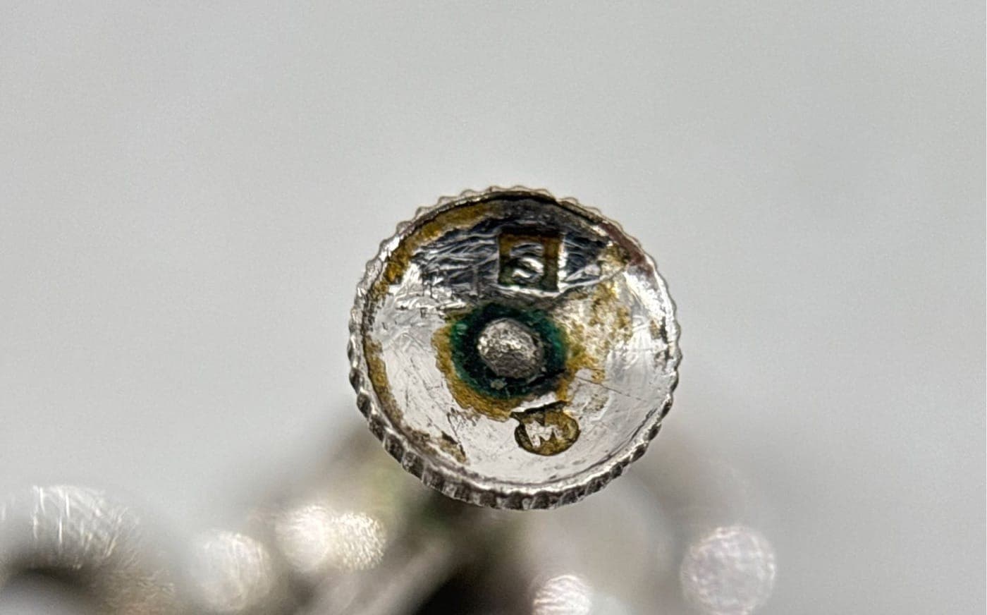 Counterfeit Mikimoto screw-back earring fitting. Heavy green corrosion rings the hallmark, and the stamp itself is an illegible blob — not the clean M-in-shield of a genuine mark.