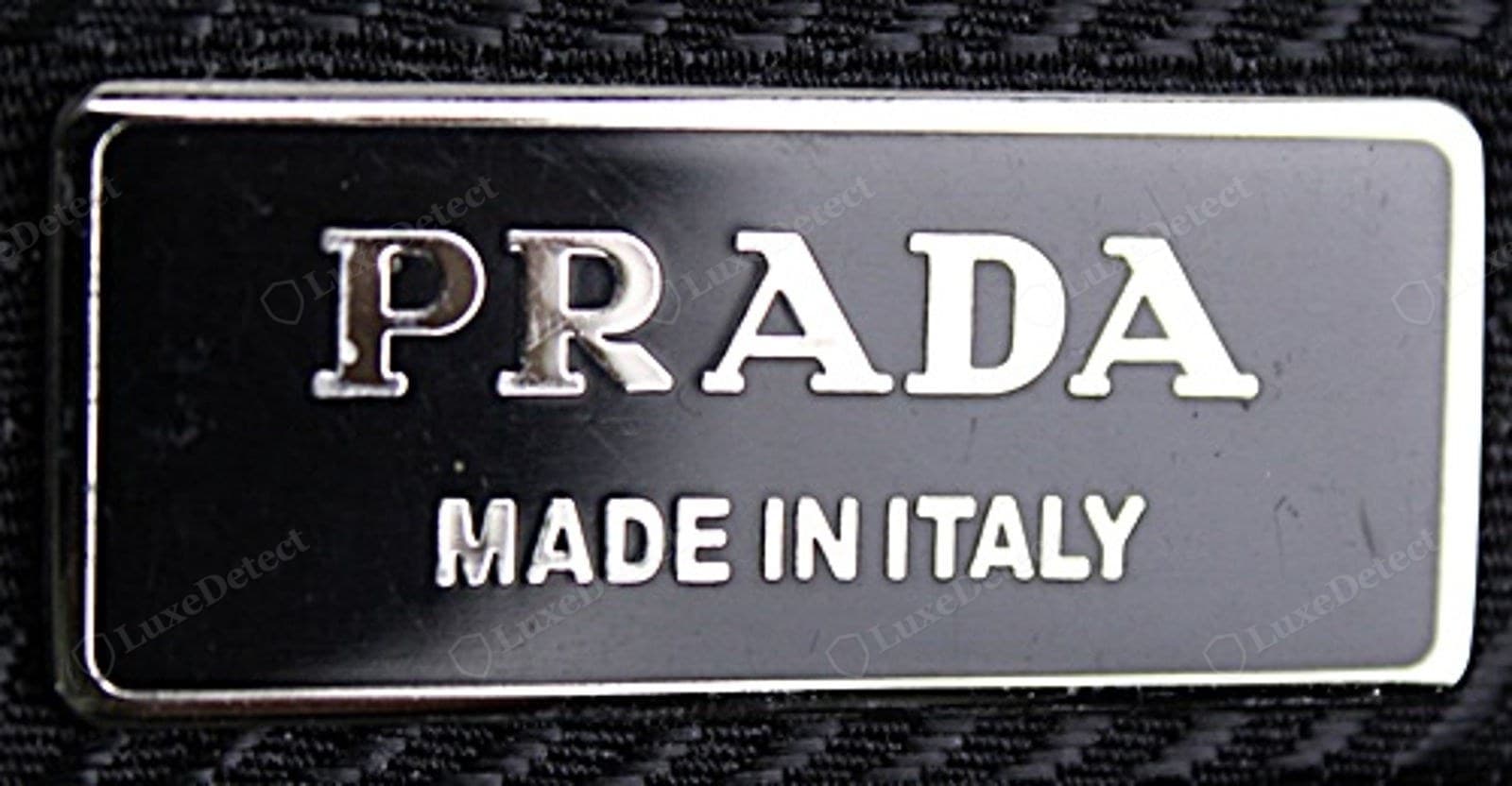 Counterfeit Prada Re-Edition metal plaque — faintly furry "PRADA" lettering, domed or matte enamel, a plaque that lifts at a corner