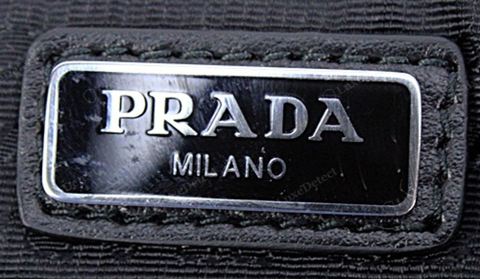 Genuine Prada Re-Edition metal triangle plaque — the "PRADA" over "MILANO" and the founding line crisp under a loupe, the enamel glossy and level