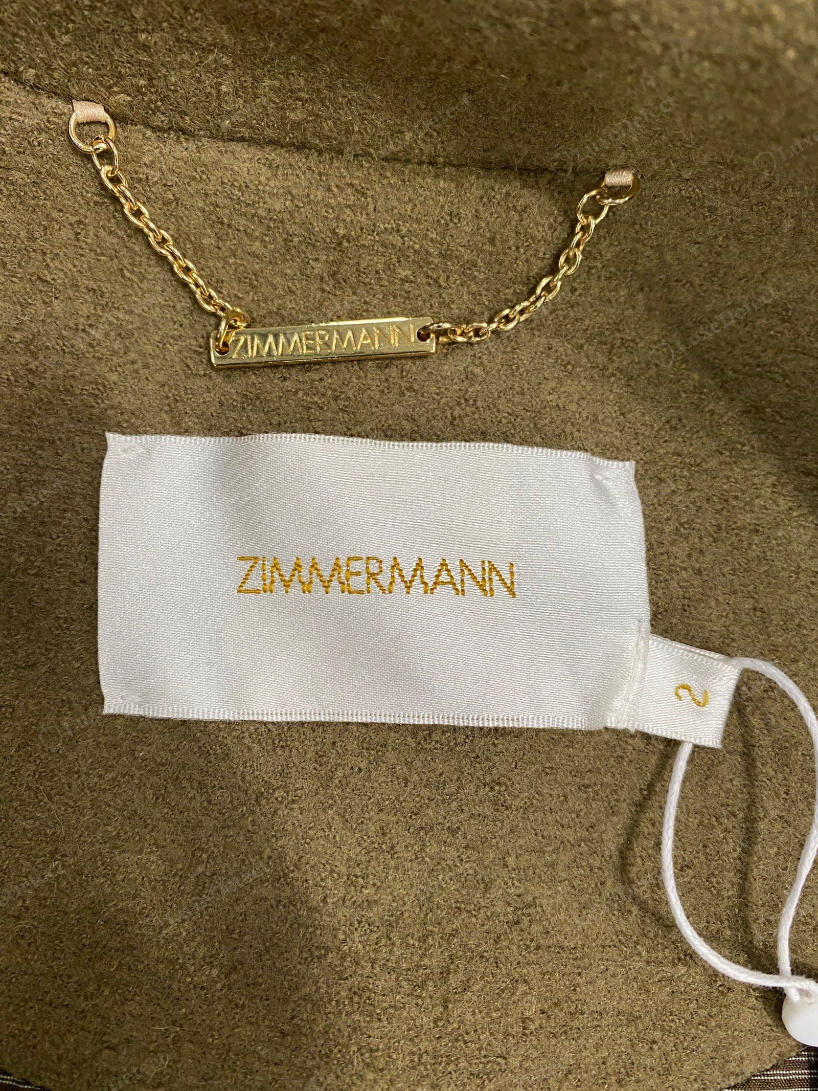 Genuine Zimmermann hanger detail — an engraved "ZIMMERMANN" gold bar plate on the hanger chain, sitting above the woven satin label and size "2" tab on an olive wool coat