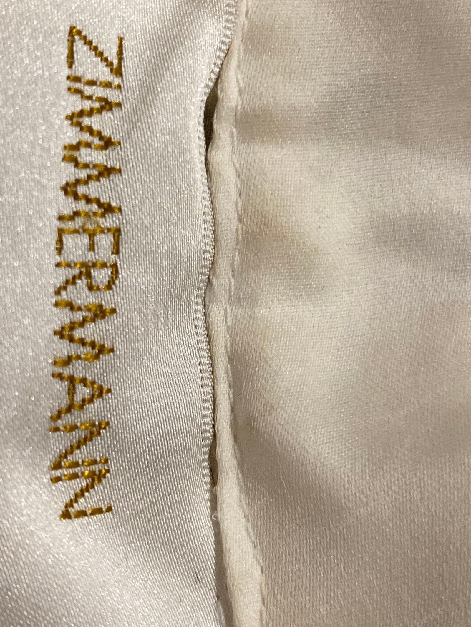Counterfeit Zimmermann label — "ZIMMERMANN" in coarse brassy gold embroidery, the stitching jagged and the letters lumpy and irregular, a second "M" malformed