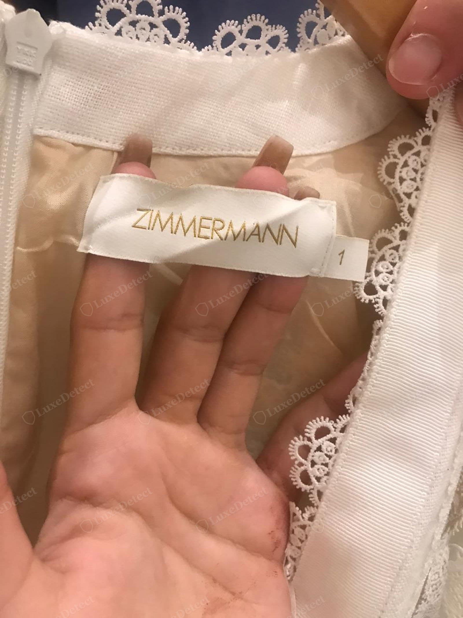 Genuine Zimmermann label — "ZIMMERMANN" in fine flat gold thread on a white satin tab, the letterforms crisp and even, with a separate woven size tab, sewn into an ivory dress with scalloped lace trim