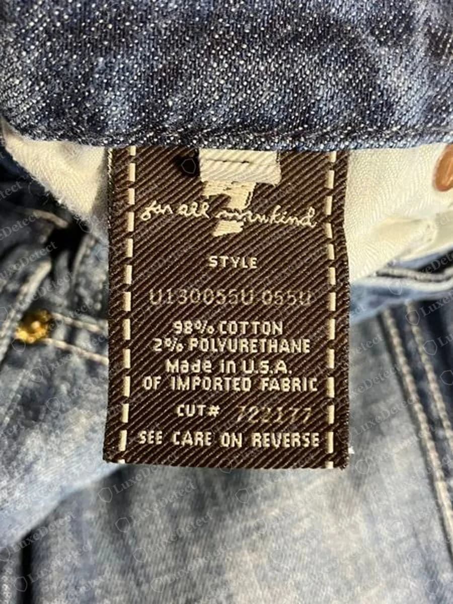 Genuine 7 For All Mankind brown twill woven waistband label with white dashed stitching, a large 7 and the script for all mankind, then style number, 98% cotton 2% polyurethane, Made in U.S.A. OF IMPORTED FABRIC, a cut number and SEE CARE ON REVERSE