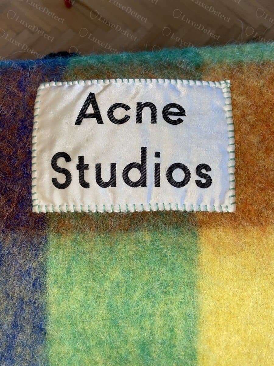 Authentique : patch logo d'angle d'une écharpe Acne Studios, avec le nom en lettres régulières et bien espacées, cousu à plat sur tout le pourtour