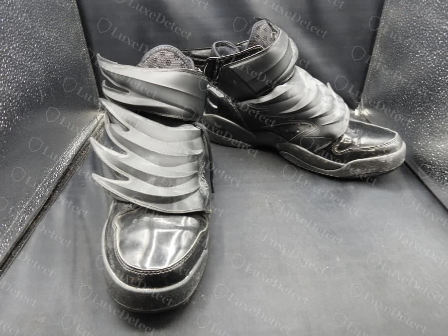 Zapatillas adidas Jeremy Scott Wings 3.0 originales, negras, sobre una silla oscura, con puntera de charol y un conjunto de alas puntiagudas grises en capas sujetas al tobillo con una correa
