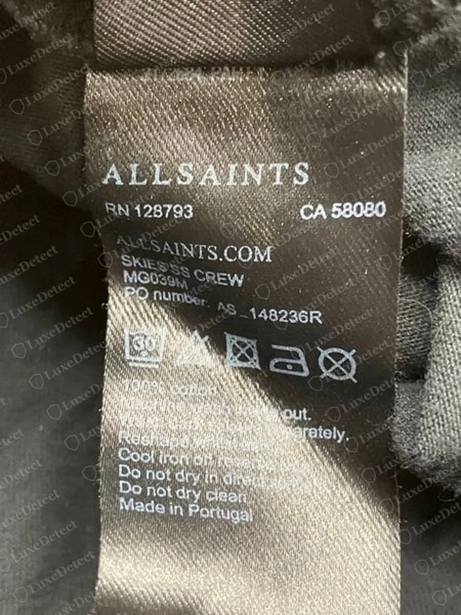 Genuine AllSaints black satin-weave care label from a Skies short-sleeve crew T-shirt showing ALLSAINTS, RN 128793, CA 58080, ALLSAINTS.COM, SKIES SS CREW, MG039M, a PO number and Made in Portugal
