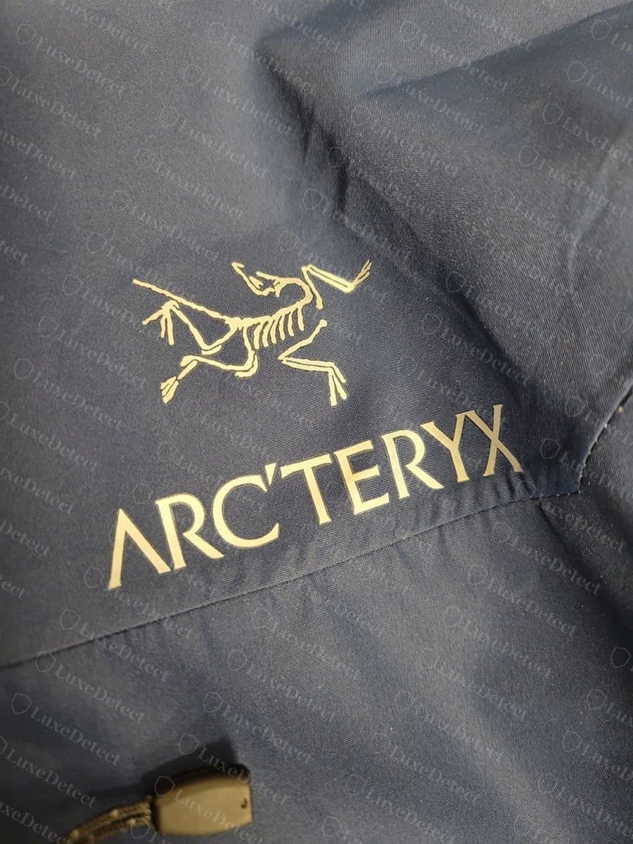 Uccello e logotipo Arc'teryx originali color oro su una giacca shell blu navy, bordi nitidi con le penne primarie distinte