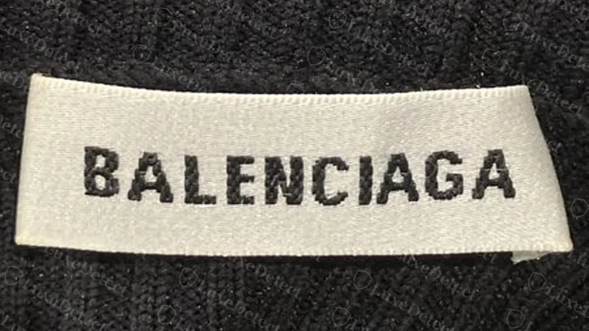 Echtes gewebtes Balenciaga-Nackenetikett auf einem schwarzen Rippstrick: cremeweißes Satinband, BALENCIAGA in schwarzen Blockbuchstaben, versäuberte Kanten