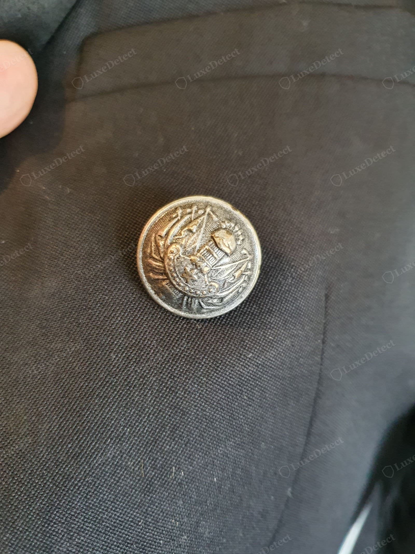 Genuine Balmain button close-up — a single antiqued gold crest button, the raised heraldic design sharp and fully defined against the black wool