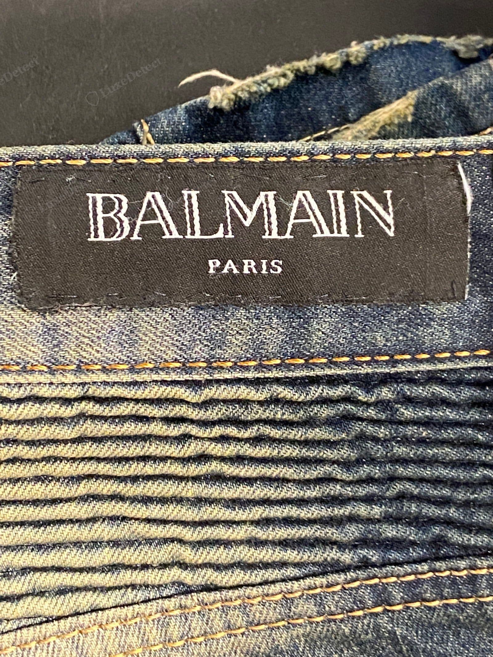 Counterfeit Balmain label — "BALMAIN PARIS" flat-printed (not woven) on a black satin tag with slightly uneven letterforms, stitched on skew with loose threads at a fraying denim seam