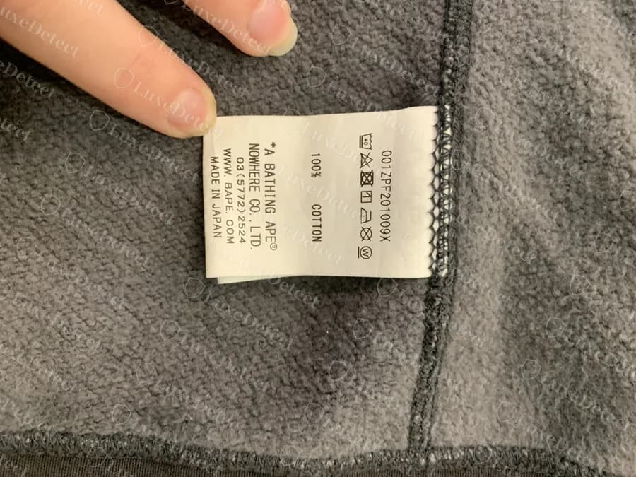 Etiqueta de cuidados original da BAPE dentro de um hoodie shark de camo preto: código de estilo, linhas “A BATHING APE” e NOWHERE CO., LTD., telefone, WWW.BAPE.COM e “MADE IN JAPAN”. Os hoodies falsos trazem este mesmo layout