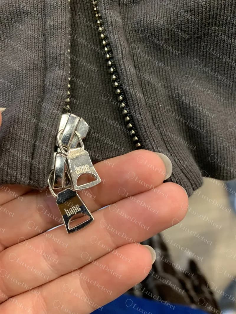 Echte BAPE-Zipper an einem Black-Camo-Shark-Hoodie: zwei silberne Metallzipper mit der Beschriftung "bape" am Schieber. Gefälschte Hoodies tragen dieselbe Beschriftung