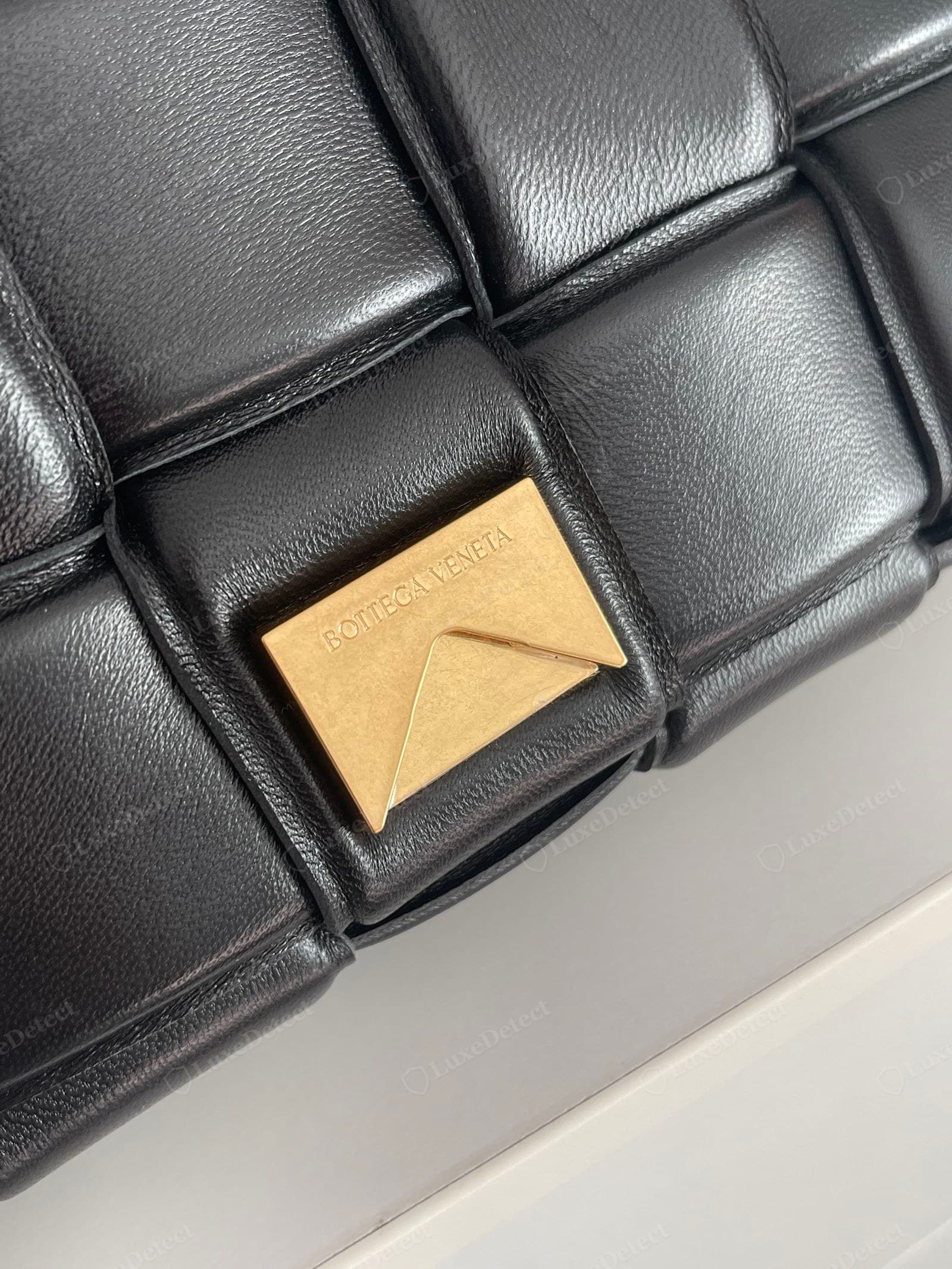 Genuine Padded Cassette flip-lock: a solid brushed antique-gold rectangular plate on a raised woven square, engraved "BOTTEGA VENETA" in fine wide-tracked serif capitals, cut crisp and centred
