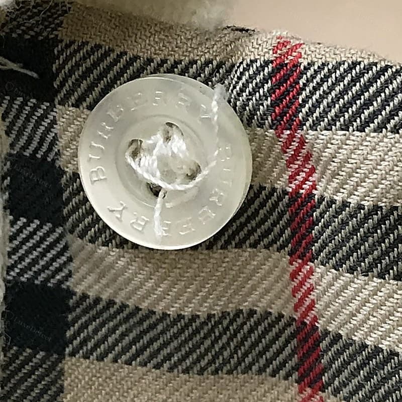 Botón Burberry original: resina color crema nacarada, "BURBERRY" grabado dos veces alrededor del borde en la tipografía con serifa de la casa, nítido y de corte parejo, cosido con una puntada en cruz prolija sobre una tapeta de cuadros