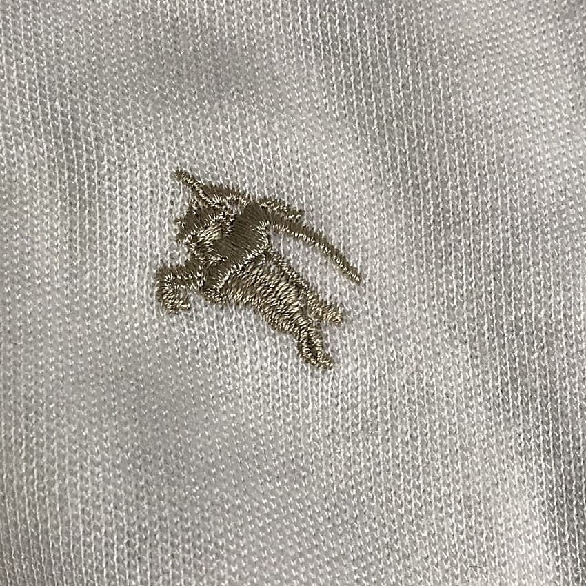 Bordado del Equestrian Knight de un Burberry falso: el caballero y el caballo reducidos a una mancha color beige abultada, las patas fundidas, la lanza fragmentada, densidad de puntadas desigual y un cabo de hilo suelto