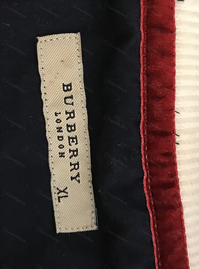 Etiqueta del cuello Burberry falsa: "BURBERRY LONDON" en una serifa condensada y ligeramente equivocada sobre un tejido más suelto, con los bordes de la etiqueta sin rematar y deshilachados, torcida respecto de la costura