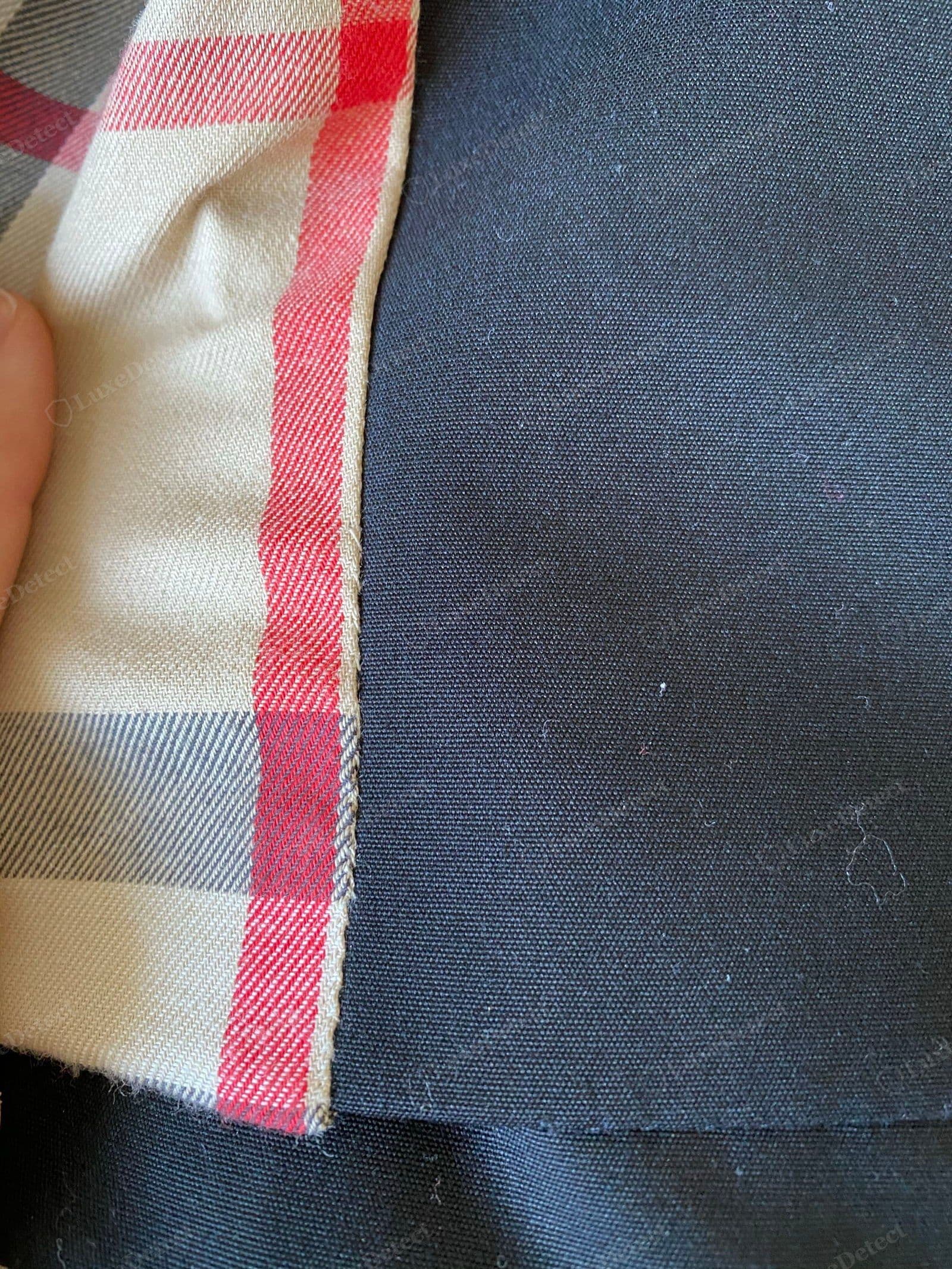 Counterfeit Burberry trench cloth — a loose, flat plain-weave shell with no diagonal twill wale, showing surface lint and pilling, against a check lining with a raw fraying edge