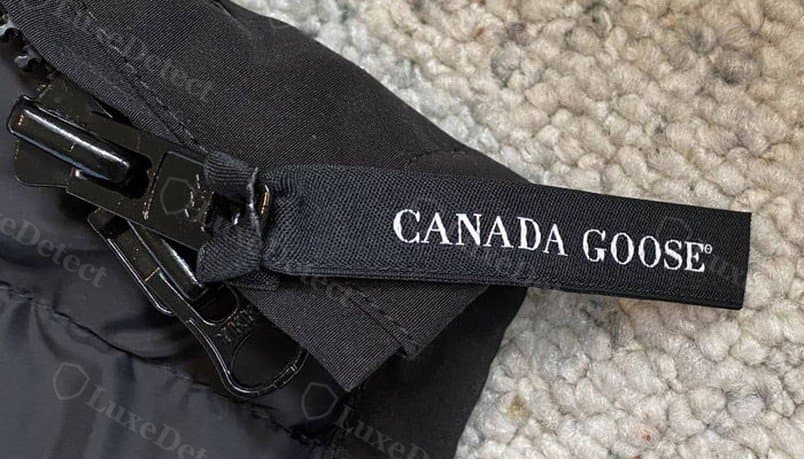 Tirador de cierre de una falsificación en un anuncio: un tirador negro liso sin ninguna marca visible del fabricante en el propio herraje, junto a una presilla de tela con la marca "CANADA GOOSE".