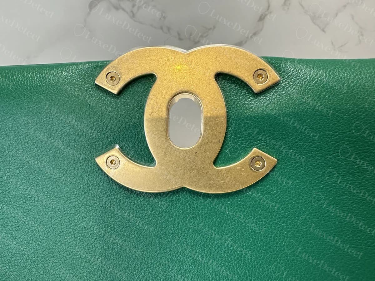 Reverso de un cierre CC original de Chanel 19: una placa CC plana en tono dorado con una abertura ovalada en el centro, sujeta con cuatro tornillos pequeños, vista en la parte inferior de la solapa