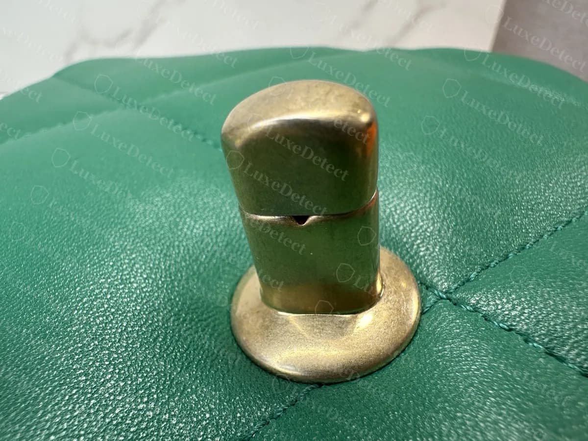 Pasador metálico en el frente del cuerpo de un Chanel 19 original verde: un cilindro redondeado en tono dorado sobre una base redonda plana, con una pequeña muesca, colocado sobre la piel acolchada