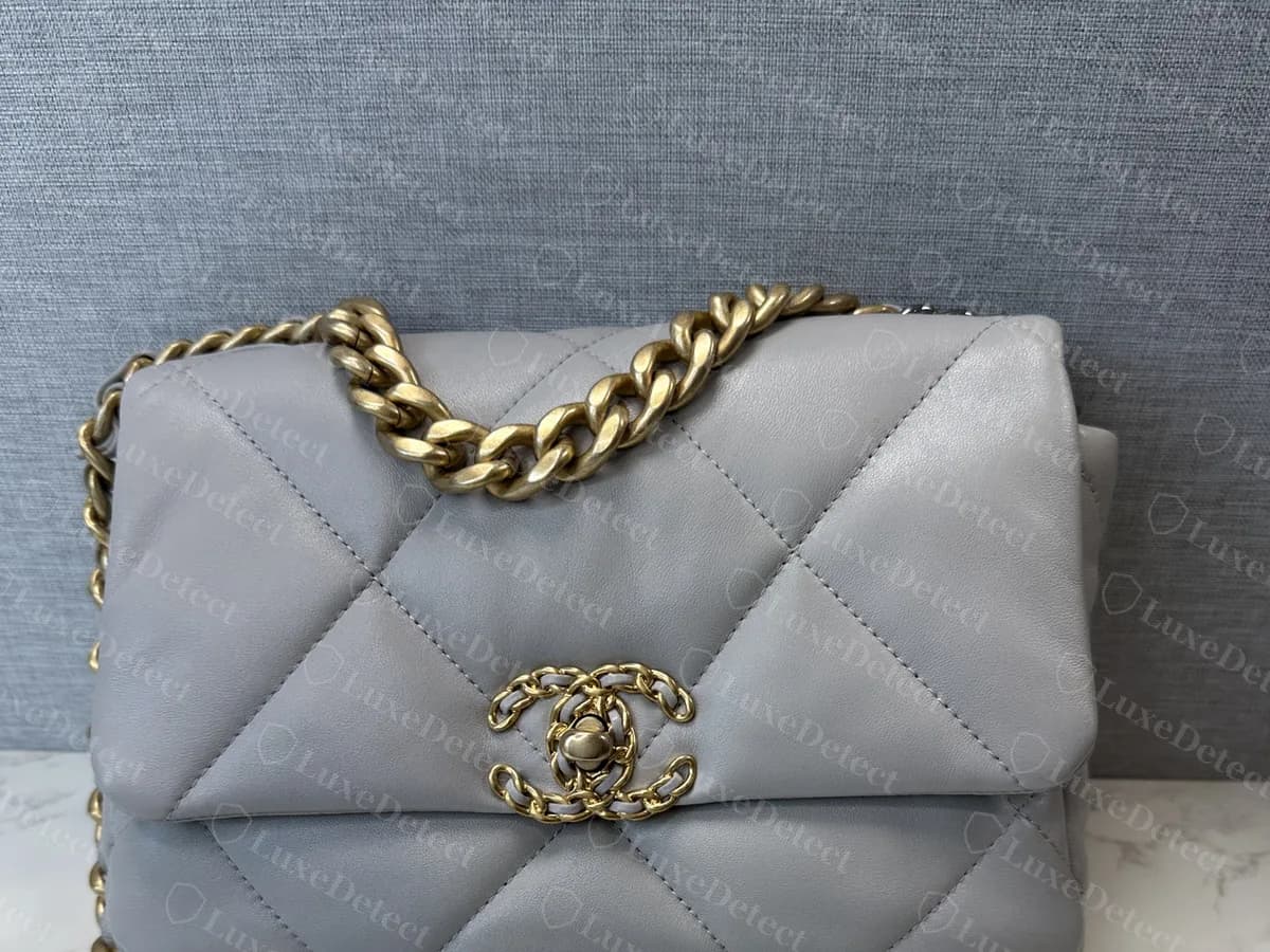 Chanel 19 flap Medium original gris visto de frente: una solapa suave y redondeada con rombos acolchados grandes, el cierre CC en el centro y una cadena dorada en la parte superior con cadena oscura colgando debajo