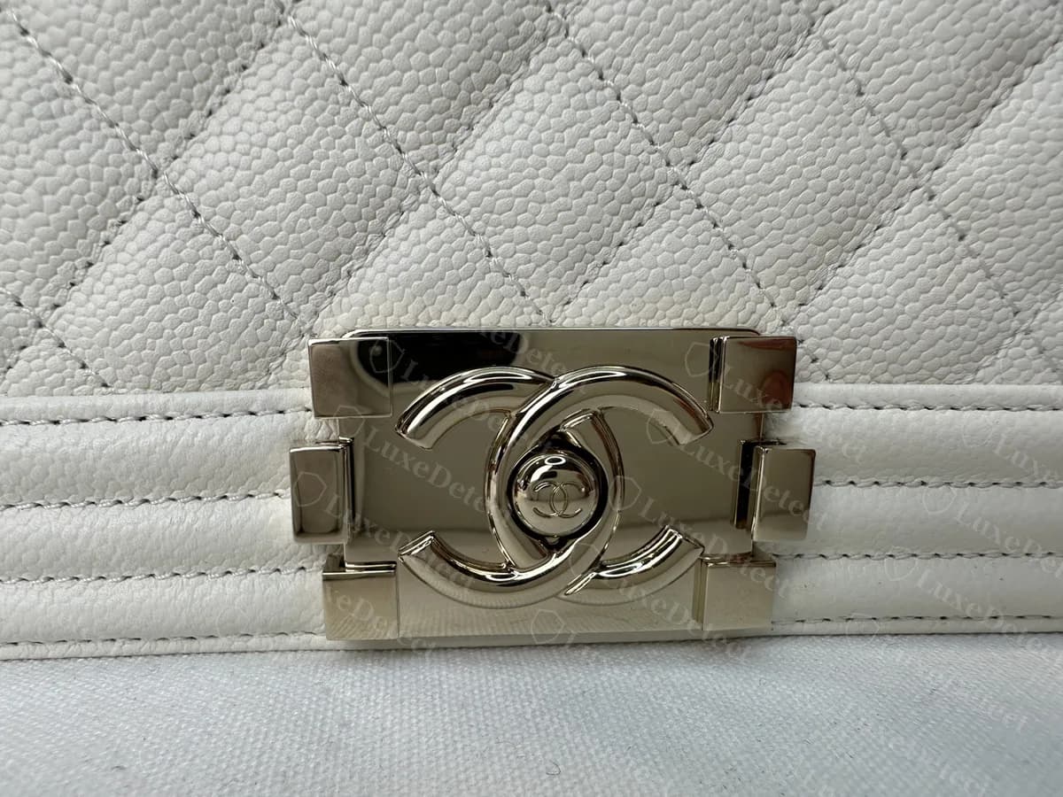 Verschluss einer echten Boy Bag in hellem Goldton: ineinandergreifendes CC um ein rundes Mittelstück auf einer rechteckigen Platte mit kleinen quadratischen Blöcken am Rand, auf weißem Caviar-Leder