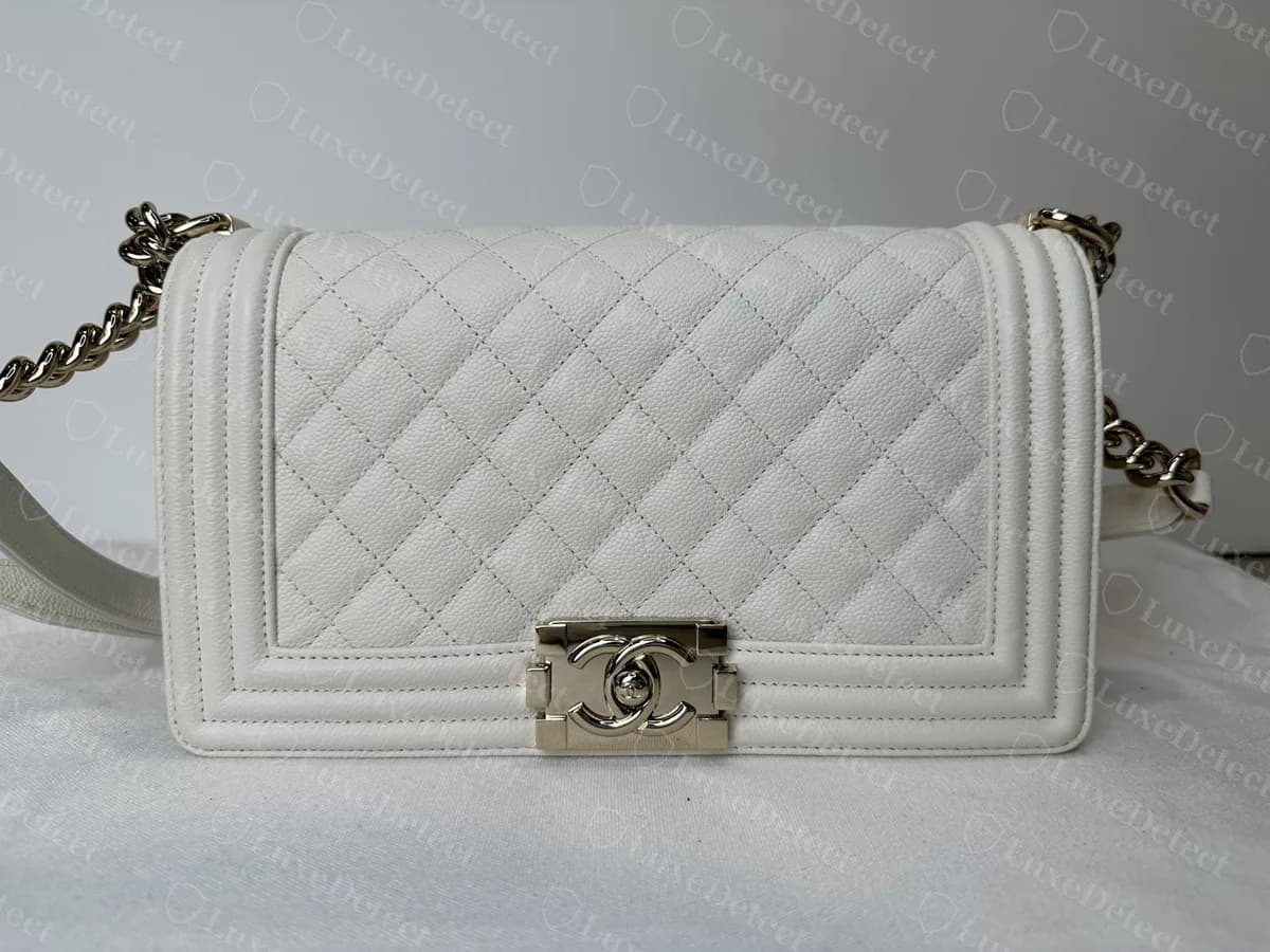 Sac Chanel Boy authentique en caviar blanc, vu de face : matelassage en losanges sur le rabat, lignes de bordure en relief tout autour, fermoir rectangulaire et chaîne dorée