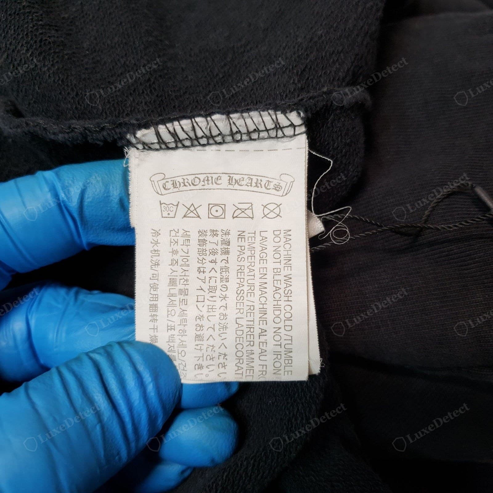 Counterfeit Chrome Hearts care label — the scroll cartouche blackletter is fuzzy and thick, and the tag is sewn in with messy overlock stitching, loose threads and a stray hang-tag string