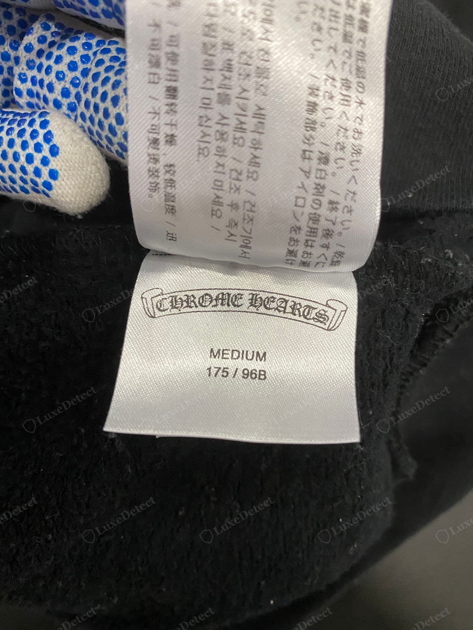 Genuine Chrome Hearts apparel care label — the "CHROME HEARTS" scroll cartouche in crisp blackletter on a white satin tab, "MEDIUM 175/96B" sizing and multilingual care text, sewn flat into black French terry