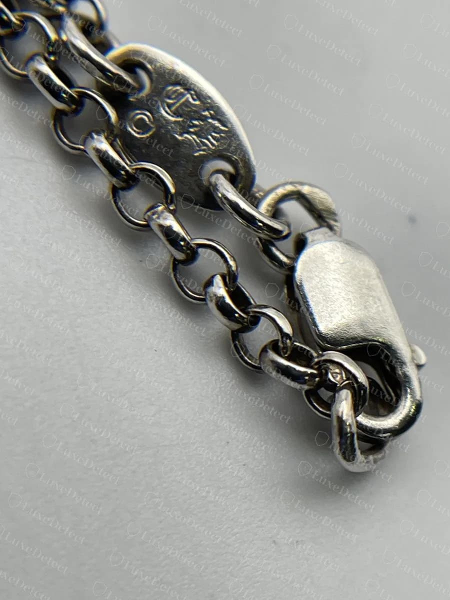 Contraffatto: l’estremità di una collana roll chain Chrome Hearts falsa, un cartellino ovale i cui marchi CH sono morbidi e a stento leggibili, accanto a un moschettone semplice e liscio