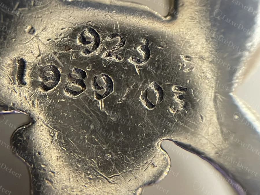 Falso: el reverso de un colgante de daga falsificado de Chrome Hearts, con un sello ".925" y un año con números de distintos tamaños, una línea base despareja y un golpe blando y poco profundo entre arañazos profundos en la superficie