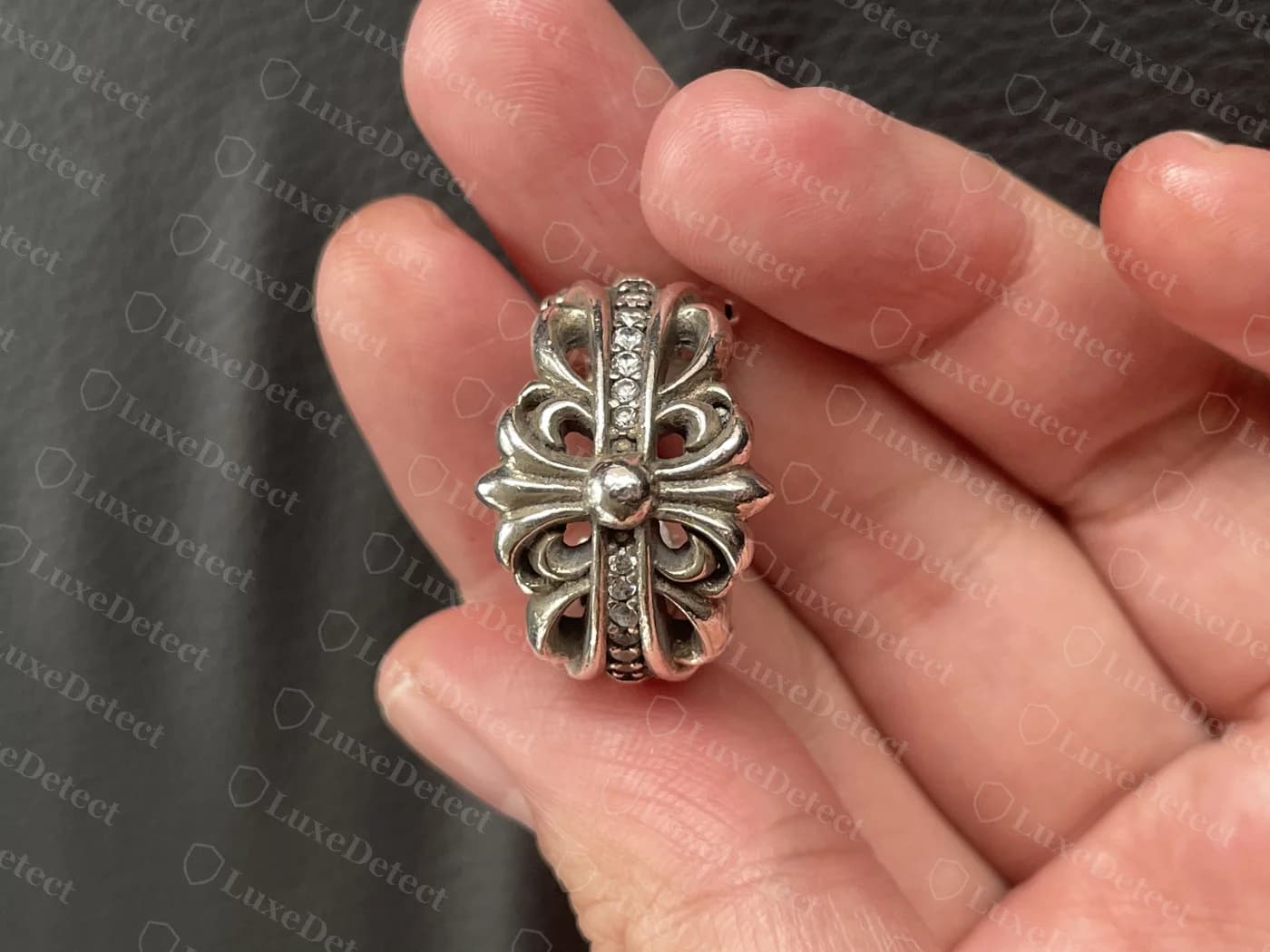 Genuine Chrome Hearts fleur-de-lis cross ring held in hand, showing deep dimensional carving with clean undercuts between the scroll elements and a pavé-set band