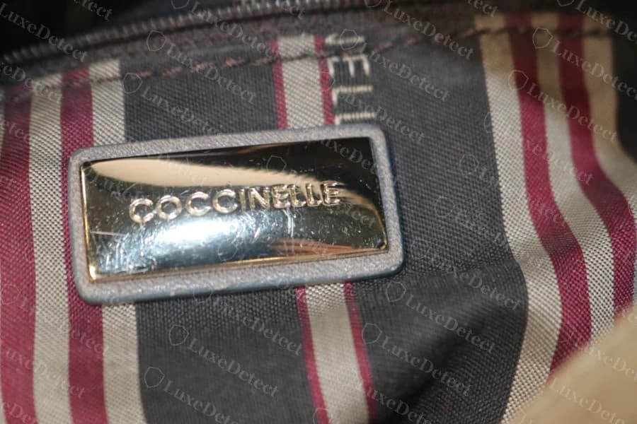 Echte gravierte goldfarbene Coccinelle-Metallplakette mit der Aufschrift COCCINELLE in einem grauen Lederrahmen, auf gestreiftem bordeauxrotem und beigem Futter