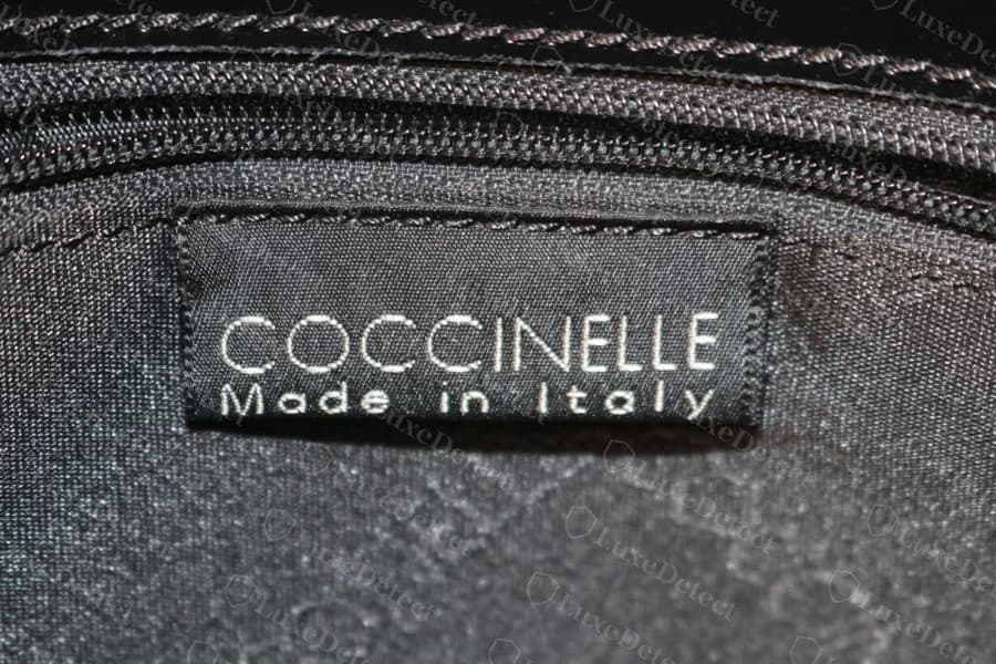 Etiqueta interna tecida Coccinelle original com COCCINELLE acima de Made in Italy, letras claras sobre etiqueta preta costurada ao forro escuro ao lado de um zíper