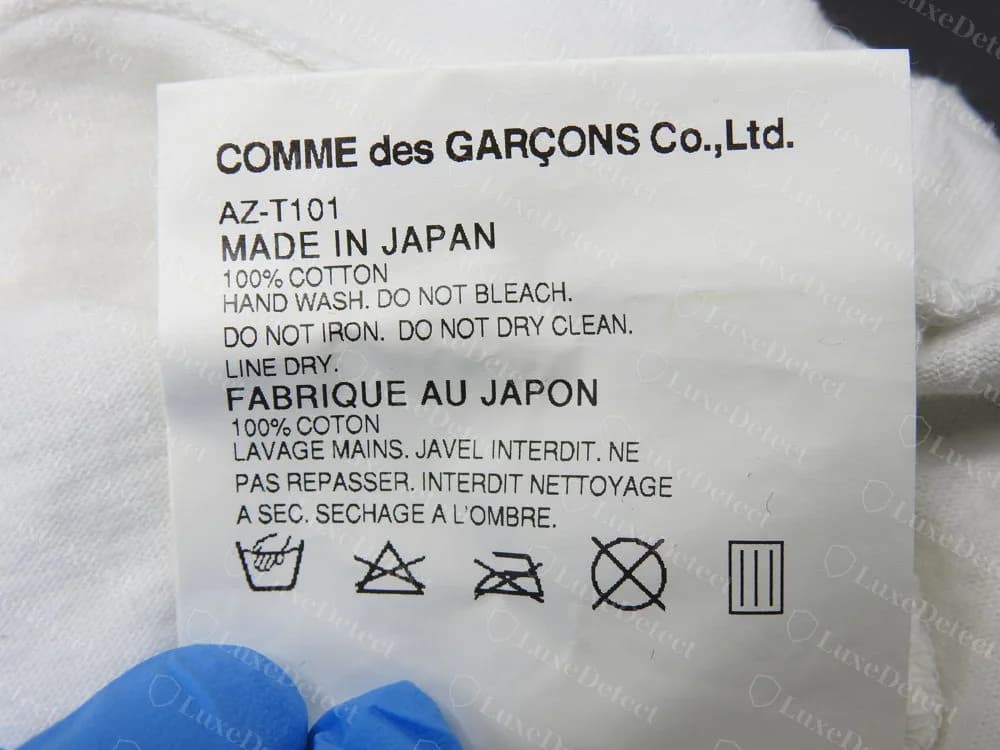 Genuine PLAY English and French care tag: COMME des GARÇONS Co.,Ltd., style code AZ-T101, MADE IN JAPAN with 100% cotton care wording, then FABRIQUE AU JAPON with the French wording and wash symbols