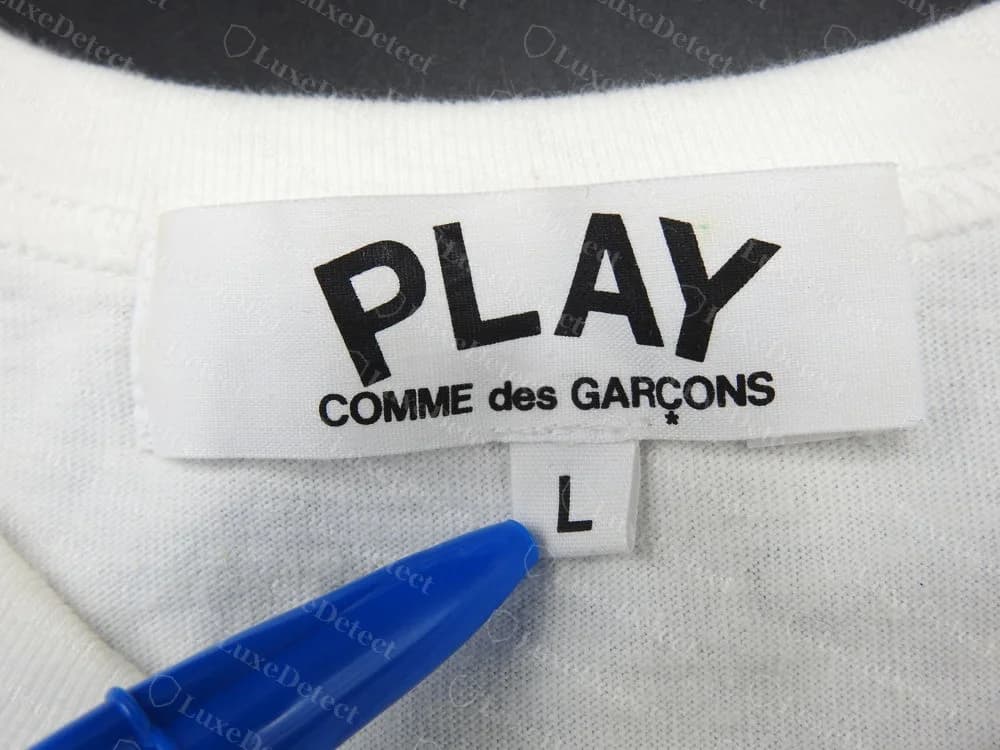 Etiqueta tejida de cuello genuina de PLAY Comme des Garçons en una camiseta blanca: PLAY en grandes mayúsculas negras sobre COMME des GARÇONS con cedilla y una pequeña estrella bajo el nombre, y una pestaña blanca pequeña y separada debajo que muestra la talla L
