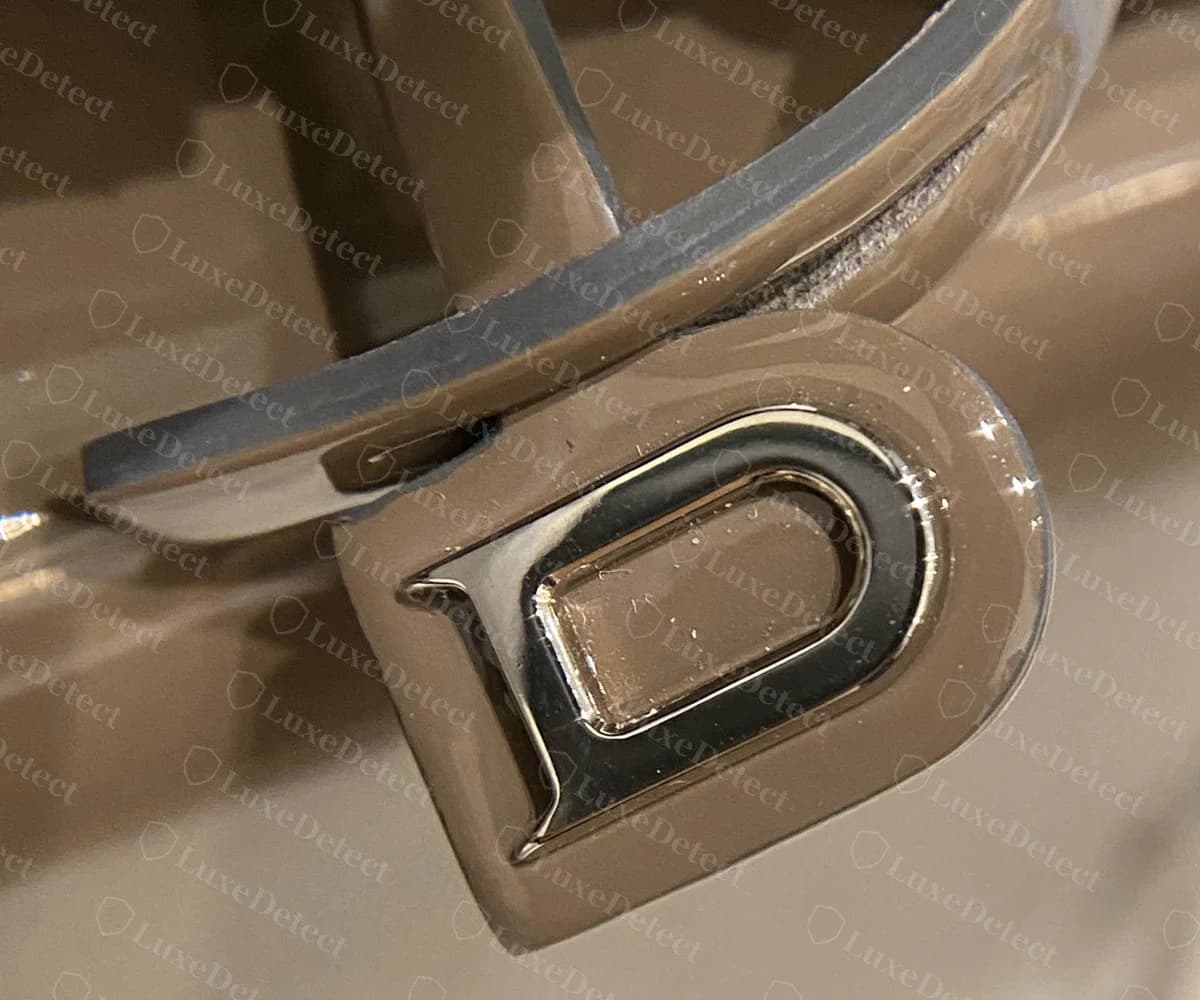 Genuine Delvaux Tempête D charm, a leather tab set with a polished silver-tone D, hanging from the handle of a taupe patent bag