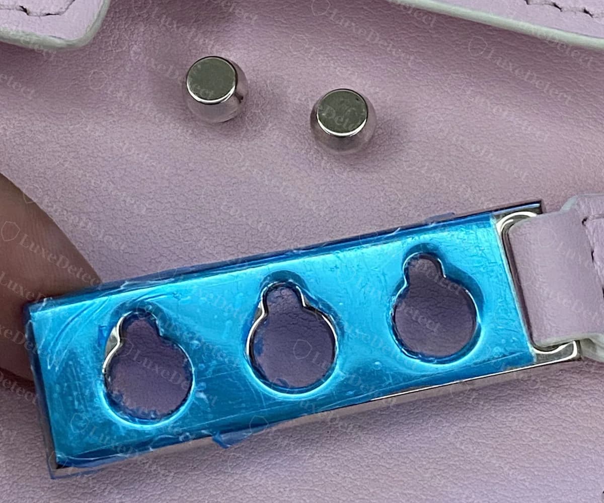 Genuine Delvaux Tempête side plate lifted off the body, showing three keyhole openings in the plate and the two round metal studs it hooks onto, with the factory protective film still on the metal