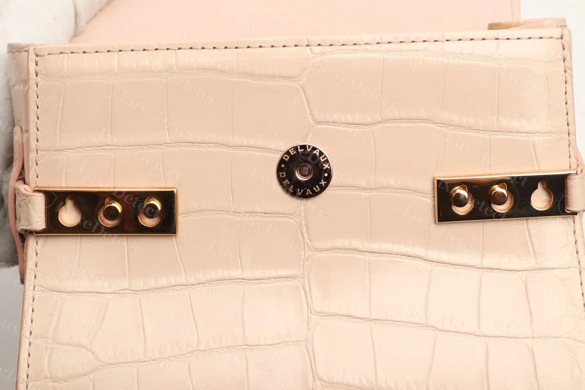 Genuine crocodile-embossed Delvaux Tempête closing with a round snap engraved DELVAUX around its rim, between the two gold-tone side plates