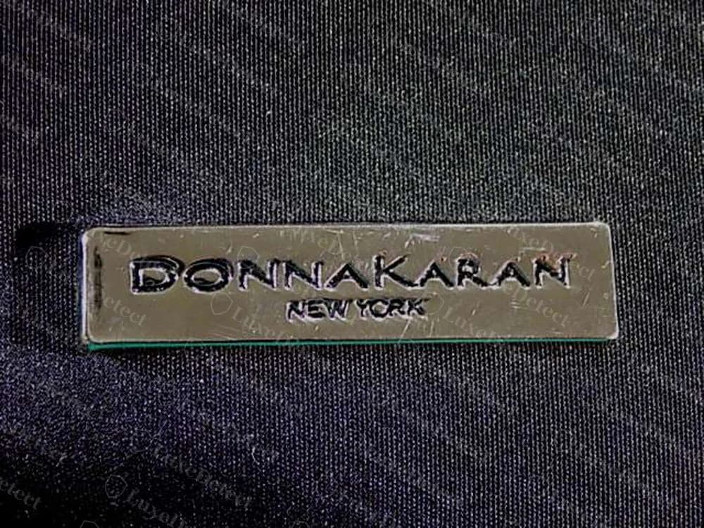 Placa de metal retangular DKNY original gravada DONNAKARAN com NEW YORK em letras menores abaixo, fixada em tecido escuro