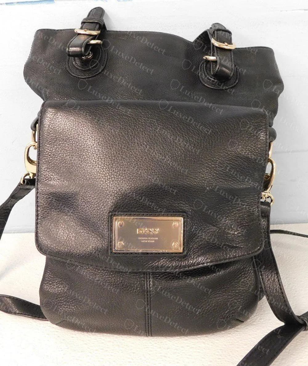 Bolso original de DKNY de solapa en piel granulada negra con una placa rectangular dorada que dice DKNY sobre una línea más pequeña de Donna Karan New York, sujeta con cuatro tornillos en las esquinas