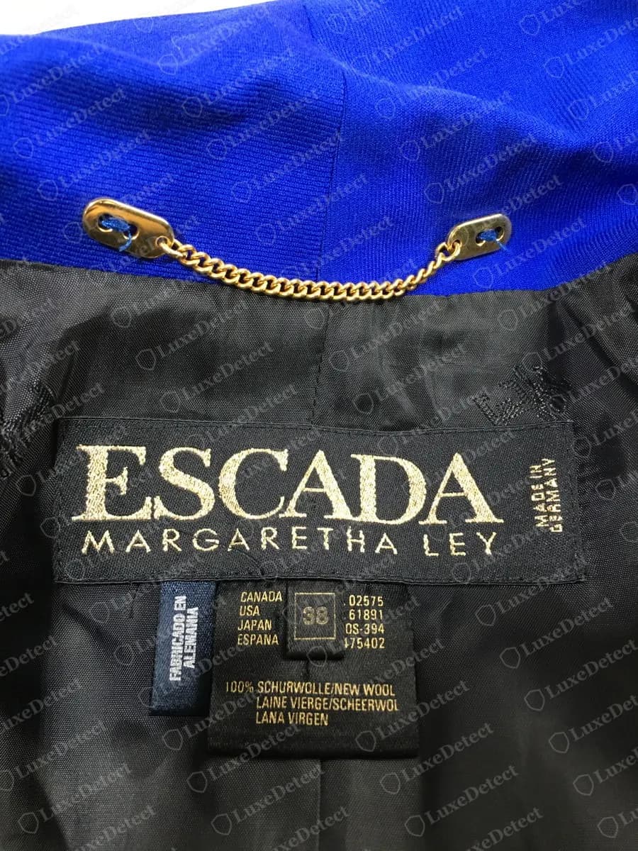Genuine Escada black woven jacket label in gold lettering: ESCADA above MARGARETHA LEY with MADE IN GERMANY running vertically on the right, and below it a navy tag reading FABRICADO EN ALEMANIA, a size box marked 38 beside CANADA, USA, JAPAN and ESPANA columns, and a fibre line for 100% new wool