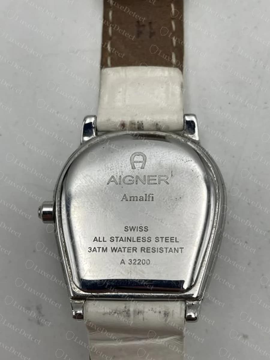 Genuine Aigner Amalfi watch caseback on a white strap, horseshoe-shaped, engraved with the A emblem, AIGNER, Amalfi, SWISS, ALL STAINLESS STEEL, 3ATM WATER RESISTANT and reference A 32200